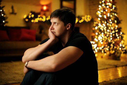 Lonely young man bored in a room decorated with a festive christmas tree and garlands on new years eve. Selective soft focus, film grain effect