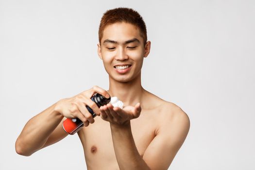 Beauty, people and hygiene concept. Portrait of handsome asian man with naked torso standing in bathroom and use shaving cream, spray foam on hand, smiling pleased, white background.