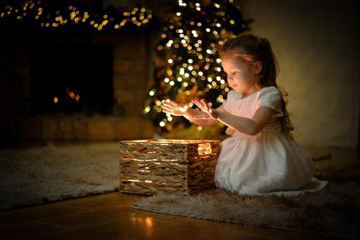 Little girl weared in white dress opened a magical New Year's gift in a Christmas interior with a Christmas tree and garlands. Selective soft focus, film grain effect
