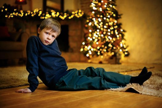 Portrait of a blond cute boy 5-7 years old sitting on the floor against the background of a festive Christmas tree. Selective soft focus, film grain effect