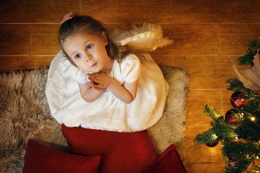 Cute blond angel girl wearing white dress 3-5 years old sitting on the carpet near the New Year tree. Top view. Selective soft focus, film grain effect
