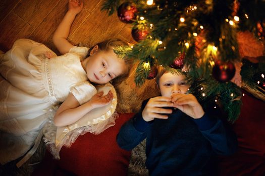 Brother and sister lies on the carpet near the New Year tree in cozy christmas interior with festive garland. Selective soft focus, film grain effect