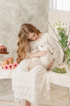 The girl is sitting at the Easter table with cakes, spring flowers and quail eggs and holding a white kid in her arms. Happy Easter celebration