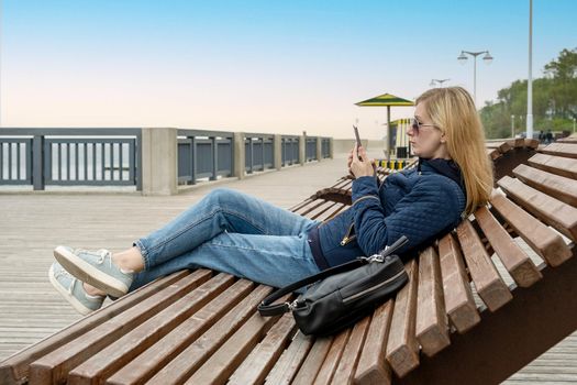 Young lonely blonde woman on empty city embankment in sun lounger uses smartphone on spring or autumn evening. Selective focus.