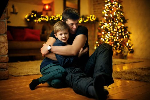 Young father hugs his son in a room decorated with a festive christmas tree and garlands on new years eve. Selective soft focus, film grain effect