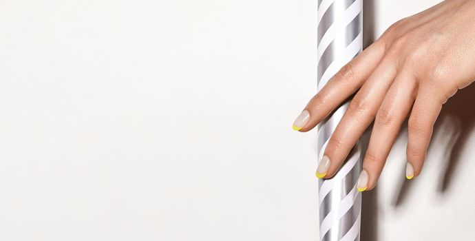 Hands with bright yellow french manicure on geometric background. Nails art design. Close-up of female hands with trendy neon nails on silver striped print tube