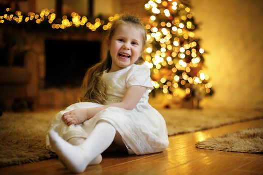 Funny little cute girl 3-5 y.o. dressed in white dress posing on floor in christmas interior with christmas tree and festive garlands. Selective soft focus, film grain effect