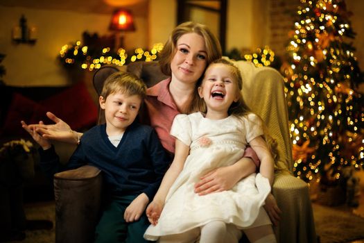 Young beautiful blonde mom with two children posing in armchair in christmas interior with a christmas tree and garlands. Selective soft focus, film grain effect