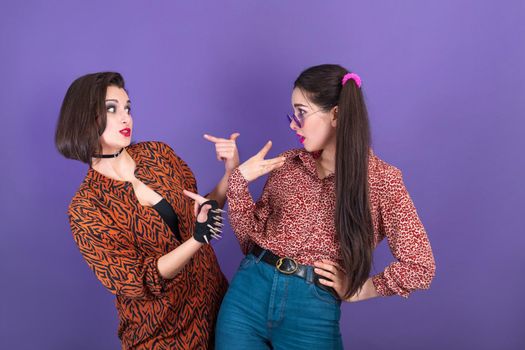 Two cute young women dressed in 90s style fooling around on purple background.