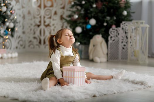 Offended little girl on a white carpet with new year gift against the background of Christmas trees.