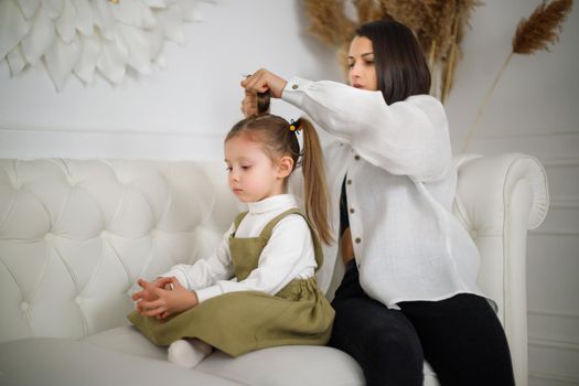 Focused mum collects daughters hair while sitting on the couch in luxury white interior.