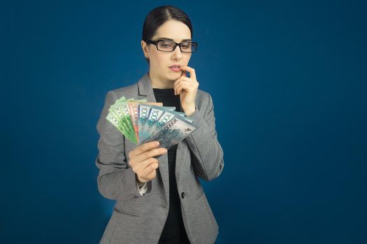 Beautiful young woman in gray jacket with tenge money in hands thought about. a blue background.