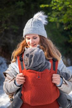 Young smiling babywearing mother with her baby in baby sling winter outdoor adventure.