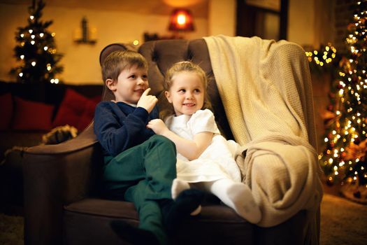 Brother and sister posing in armchair in cozy christmas interior with christmas tree and festive garland. Selective soft focus, film grain effect
