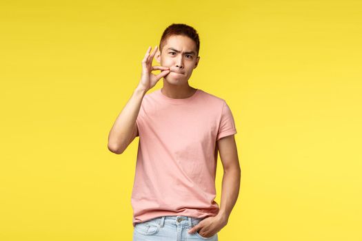 You better shut your mouth. Portrait of annoyed and serious young asian man show zip on lips, seal them as prohibit not tell anyone secret, demand silence, standing bothered yellow background.