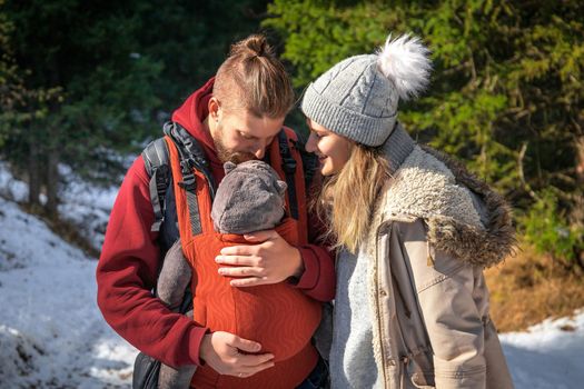 Young caucasian babywearing family with their baby in baby carrier winter outdoor adventure.