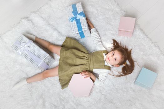Cute little girl lies on a white carpet with Christmas gifts around.