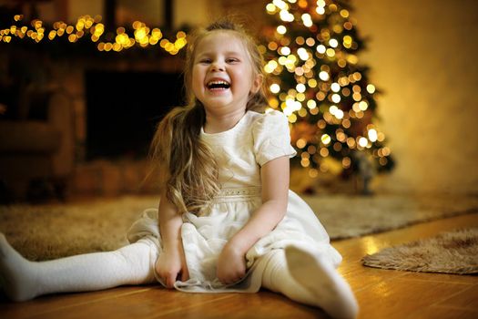 Funny little cute laughing girl 3-5 y.o. dressed in white dress posing on floor in christmas interior with christmas tree and festive garlands. Selective soft focus, film grain effect