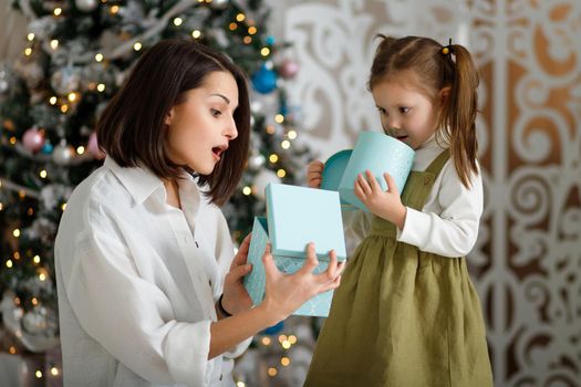 Cute little girl with her mother opening a magical Christmas gifts by a Christmas tree in cozy living room.