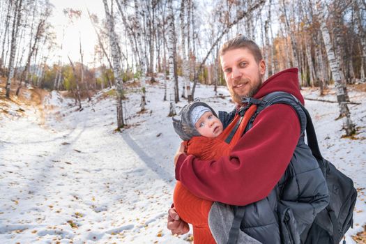 Adorable smiling bearded babywearing father with his baby in sling autumn forest walking.