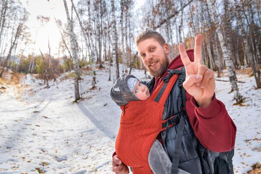 Babywearing winter forest walk with father and his baby son, peace gesture.