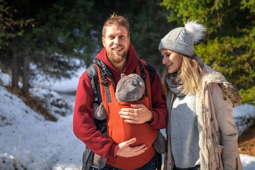 Babywearing winter forest walk with family and their baby son in ergonomic carrier.