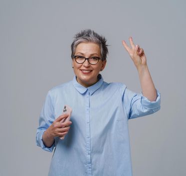 Grey haired gorgeous mature businesswoman with smartphone in hand looking at camera working or shopping online or checking on social media. Pretty woman in blue shirt isolated on white. Toned image.