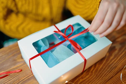 Girl in orange knitted sweater opens festive gift. Box with red ribbon on wooden table. Christmas or Birthday.