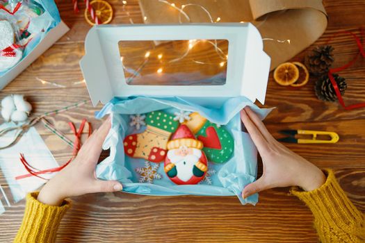Girl's hands open gift box with Christmas cookies. Gingerbread with icing. Festive sweets for children in form of santa claus, mittens, socks and snowflakes. New Year's composition on wooden table.