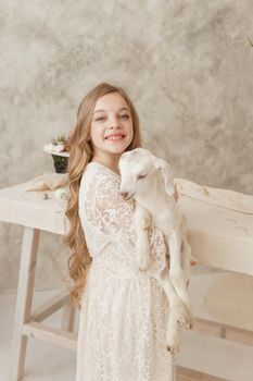 The girl is sitting at the Easter table with cakes, spring flowers and quail eggs and holding a white kid in her arms. Happy Easter celebration