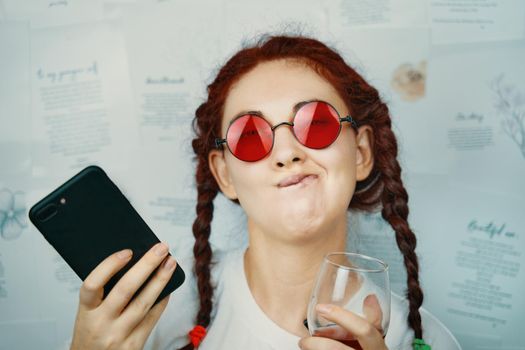 Funny girl with pigtails and red sunglasses makes faces. Female blogger with a glass of wine and a smartphone in her hand. Pampering and antics. Hippie or boho style fashion blog.