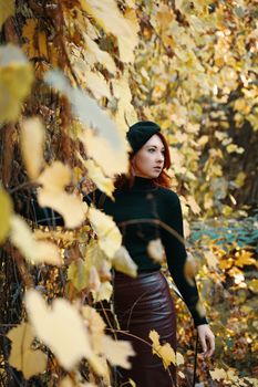 Awesome red-haired woman in autumn park. Beautiful female in black beret, knitted sweater and leather skirt. Stylish model among yellow dry foliage of trees. Walk in fall nature.