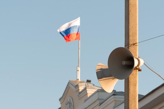 loudspeakers for emergency messages on the electrification pole. in the background of the administrative building the Russian flag is flying against the blue clear sky