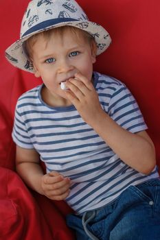 A little boy in a striped shirt is sitting on a red armchair and enjoying candy.