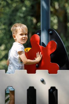 A child is driving the red helm of a ship on a playground. Dark green park tree foliage background.