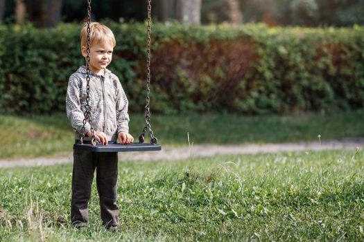 An attractive little boy stands next to a swing with chains in a city park, a child getting ready to swing. Horizontal photo.