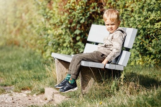 A smiling little boy sits on a wooden gray bench in a city park against the backdrop of green summer foliage and sunlight.