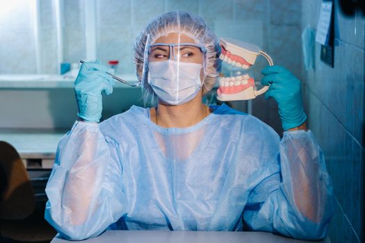 A dental doctor wearing blue gloves and a mask holds a dental model of the upper and lower jaws and a dental mirror.