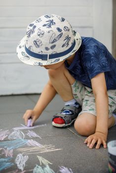 Small caucasian boy three years old sitting on the concrete pavement drawing with chalk in park in day childhood and growing up education concept