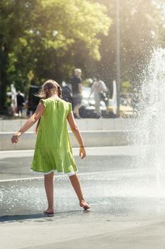 A young girl in a light green dress barefoot dances by a fountain in the city center.