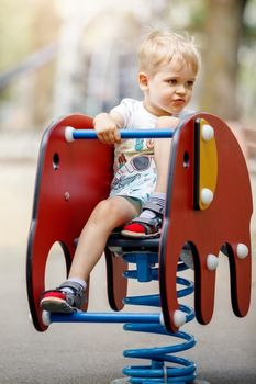 The blond little boy swings on a springy elephant-shaped swing on the playground during the summer.