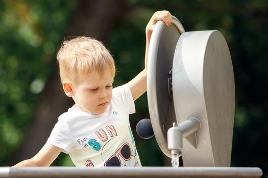 A happy little boy plays with a water tap in a city park. Special water equipment for children's games on a hot summer day outdoors.