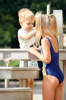 A happy boy and a cute girl in a blue bathing suit play with a water tap in a city park. Vertical.