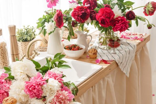 The kitchen countertop is decorated with peonies. The interior is decorated with spring flowers. Pink peonies and sweet cupcakes on a wooden countertop. Interior details.