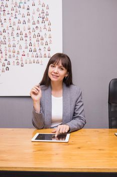 A brunette businesswoman in a gray jacket at her desk with a tablet in her hands. Business portrait in the office.