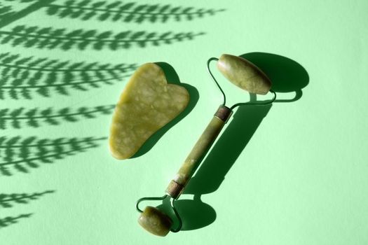 Jade Gua sha scraper and facial massager on green background. Hard light, shadows, the concept self-care. Facial care. Zero waste. Lifting and toning treatment at home.