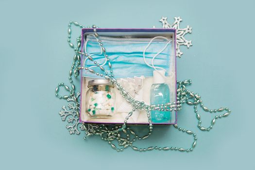 Medical mask, pills in a jar, antiseptic gel for hands and Christmas decorations in a box on a blue background. Christmas gifts in the age of coronavirus. Copy space.