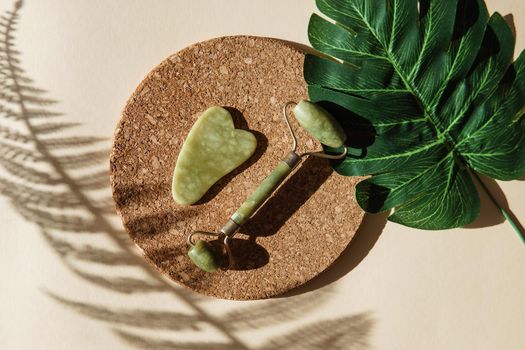 Jade Gua sha scraper and face roller massager on a cork round stand with a monstera leaf. Hard light, shadows, the concept self-care. Facial care. Zero waste. Lifting and toning treatment at home.