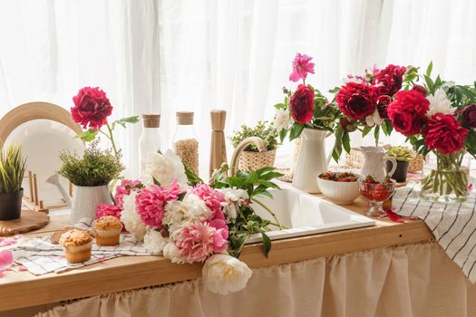 The kitchen countertop is decorated with peonies. The interior is decorated with spring flowers. Pink peonies and sweet cupcakes on a wooden countertop. Interior details