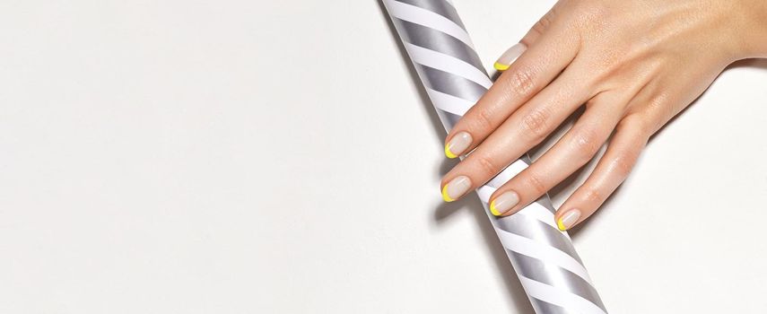 Hands with bright yellow french manicure on geometric background. Nails art design. Close-up of female hands with trendy neon nails on silver striped print tube. Copy space